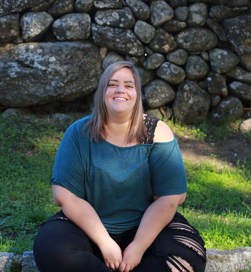 Fotografía de Cristina Castro Martínez. Mujer con una pared de piedra con musgo de fondo. Sonríe ampliamente.